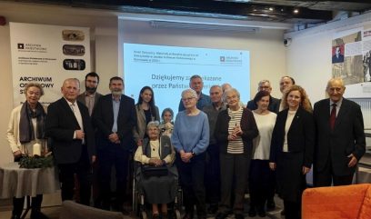 Grupa ludzi, w tym osoby starsze, stoją razem w pomieszczeniu i uśmiechają się do kamery. Niektórzy stoją, a jedna kobieta siedzi na wózku inwalidzkim. Za nimi na ekranie wyświetlany jest polski tekst. Na ścianach widoczne są archiwalne plakaty.