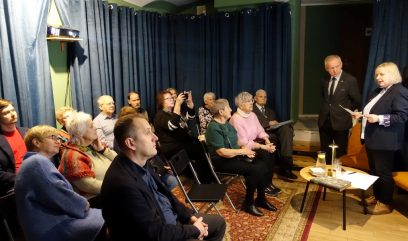 Mała grupa dorosłych siedzi na składanych krzesłach w przytulnym, zasłoniętym pokoju, słuchając kobiety stojącej i czytającej z gazet. Obok niej stoi mężczyzna w garniturze. Na stołach stoją świece, napoje i ciastka. Ludzie są uważni; jedna osoba robi zdjęcie.