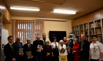 Grupa osób, głównie starszych, pozuje w pomieszczeniu przypominającym bibliotekę. Niektórzy trzymają certyfikaty. Młoda dziewczyna stoi z przodu trzymając rysunek. Za grupą stoi udekorowana choinka, a regały z książkami ustawione są wzdłuż prawej ściany. Wszyscy patrzą w stronę kamery.