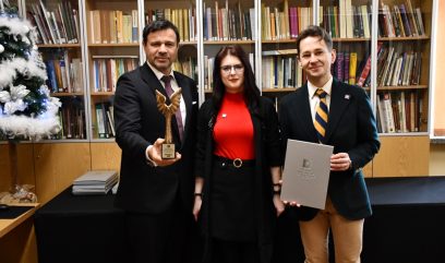 Trzy osoby stoją przed półkami z książkami. Mężczyzna po lewej, w czarnym garniturze, trzyma skrzydlate trofeum. Kobieta w środku nosi okulary i czerwoną bluzkę z czarną spódnicą. Mężczyzna po prawej, w ciemnej marynarce, trzyma teczkę. Udekorowana choinka jest widoczna po lewej stronie.