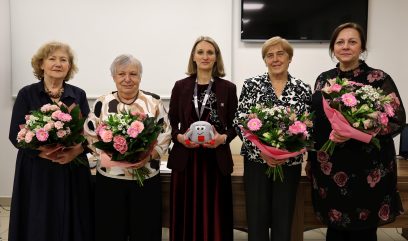 Pięć kobiet stoi w pomieszczeniu, cztery trzymają bukiety różowych kwiatów, a jedna w centrum trzyma pluszową zabawkę w kształcie uśmiechniętego zęba. Są ubrane w formalne stroje, pozują i uśmiechają się przed pustą białą ścianą i dużym czarnym ekranem telewizora.