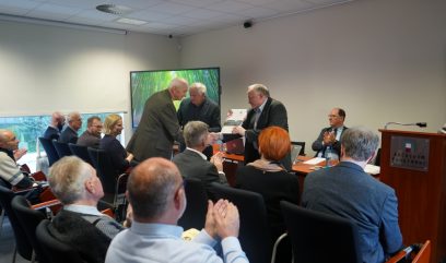 W sali konferencyjnej grupa starszych osób siedzi i bije brawo, podczas gdy trzech mężczyzn stoi z przodu. Jeden z nich wręcza drugiemu dokument lub nagrodę. Osoba przy stole przygląda się temu, a w tle widoczny jest ekran przedstawiający zielony las.