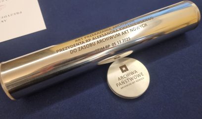 Błyszczący srebrny cylinder z wygrawerowanym polskim tekstem spoczywa na niebieskiej powierzchni. Obok znajduje się okrągła metalowa nasadka z napisem 