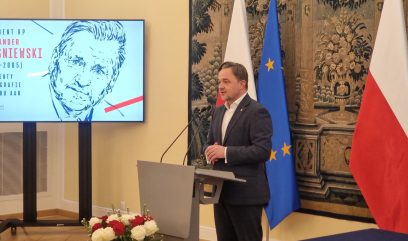 Mężczyzna w garniturze przemawia na podium. Za nim znajdują się flagi Polski i Unii Europejskiej oraz kompozycja kwiatowa. Po lewej stronie ekranu wyświetlany jest czarno-biały szkic twarzy mężczyzny, polski tekst oraz czerwone i niebieskie akcenty.