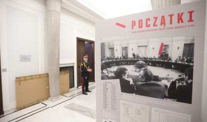 Umundurowany strażnik stoi przy kolumnie w jasnym korytarzu z marmurową posadzką. Na pierwszym planie tablica zatytułowana 