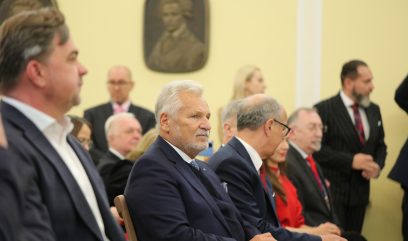 Grupa formalnie ubranych osób, głównie starszych mężczyzn w garniturach i krawatach, siedzi w rzędach na imprezie wewnętrznej. Dwie tablice z portretami wiszą na beżowej ścianie w tle. Uwaga skupiona jest na białowłosym mężczyźnie w pierwszym rzędzie.