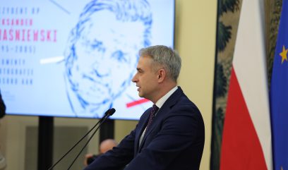 Mężczyzna w garniturze przemawia na podium. Za nim znajduje się flaga Polski, flaga UE i ekran wyświetlający niebieski szkic twarzy mężczyzny z czerwonym tekstem w języku polskim. Tło sugeruje oficjalne wydarzenie lub ceremonię.