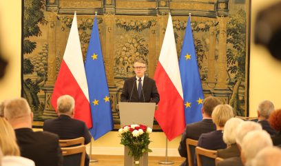 Mężczyzna w garniturze przemawia na podium przed dwiema flagami Polski i dwiema flagami Unii Europejskiej, zwracając się do publiczności siedzącej w rzędach. U podstawy podium znajduje się bukiet kwiatów, a tło zdobi duży, ozdobny gobelin.