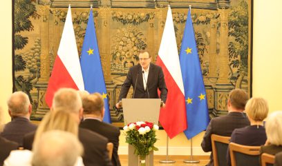 Mężczyzna w garniturze przemawia na podium przed publicznością. Za nim znajdują się dwie flagi Polski i dwie flagi UE. Ścianę zdobi duży gobelin. Bukiet czerwonych i białych kwiatów jest umieszczony przed podium w jasno oświetlonym pomieszczeniu.