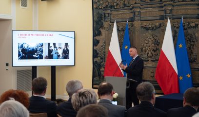 Mężczyzna w garniturze, zidentyfikowany jako Aleksander Kwaśniewski, przemawia na podium w oficjalnym pomieszczeniu z flagami Polski i UE za nim. Gestykuluje w kierunku dużego ekranu, na którym wyświetlane są historyczne zdjęcia i polski tekst; uczestnicy oglądają go pośród ozdobnych dekoracji i gobelinu.