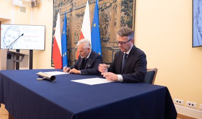 Dwóch mężczyzn w garniturach siedzi przy niebieskim stole i podpisuje dokumenty. Za nimi znajdują się flagi Polski i Unii Europejskiej, gobelin na ścianie i ekran z oficjalnym logo przedstawiającym profil Aleksandra Kwaśniewskiego w tym formalnym, oficjalnym otoczeniu.