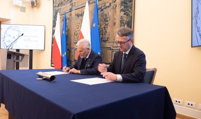 Dwóch mężczyzn w garniturach siedzi przy niebieskim stole, podpisując dokumenty. Za nimi znajdują się flagi Polski i UE oraz duży gobelin. Ekran prezentacyjny i telewizor wyświetlają treści graficzne po obu stronach. Otoczenie wydaje się formalne, prawdopodobnie jest to oficjalna ceremonia podpisania lub zawarcia umowy.