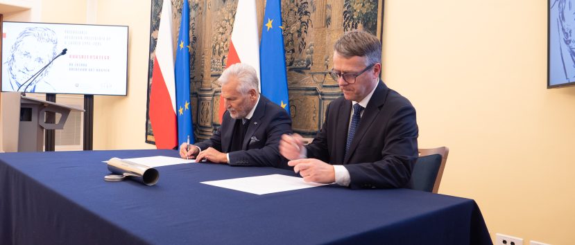 Dwóch mężczyzn w garniturach siedzi przy niebieskim stole, podpisując dokumenty. Za nimi znajdują się flagi Polski i UE oraz duży gobelin. Ekran prezentacyjny i telewizor wyświetlają treści graficzne po obu stronach. Otoczenie wydaje się formalne, prawdopodobnie jest to oficjalna ceremonia podpisania lub zawarcia umowy.