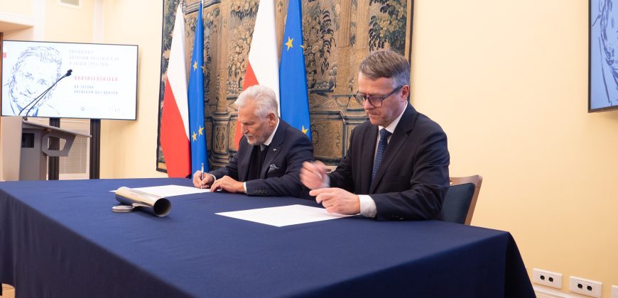 Dwóch mężczyzn w garniturach siedzi przy niebieskim stole, podpisując dokumenty. Za nimi znajdują się flagi Polski i UE oraz duży gobelin. Ekran prezentacyjny i telewizor wyświetlają treści graficzne po obu stronach. Otoczenie wydaje się formalne, prawdopodobnie jest to oficjalna ceremonia podpisania lub zawarcia umowy.