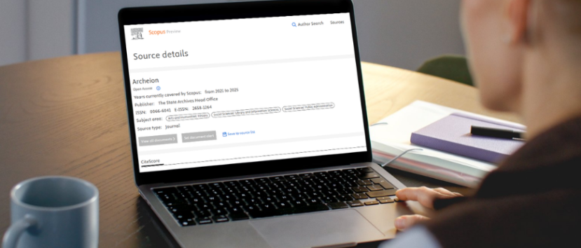 Osoba siedzi przy biurku i korzysta z laptopa. Na ekranie wyświetlana jest strona szczegółów źródła dla czasopisma Actahein w witrynie Scopus. W tle znajduje się notatnik, długopis i kubek, co wskazuje na środowisko pracy lub nauki.