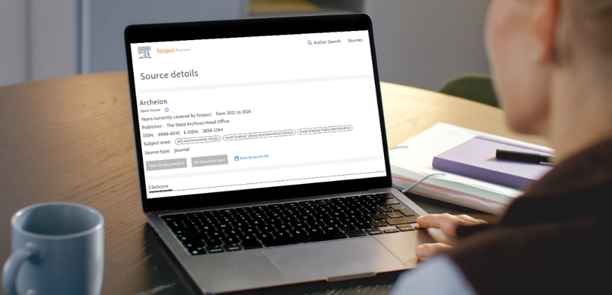 Osoba siedzi przy biurku i korzysta z laptopa. Na ekranie wyświetlana jest strona szczegółów źródła dla czasopisma Actahein w witrynie Scopus. W tle znajduje się notatnik, długopis i kubek, co wskazuje na środowisko pracy lub nauki.