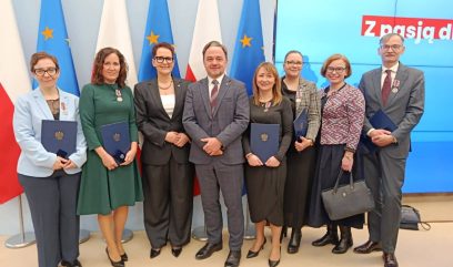 Ośmiu dorosłych, pięć kobiet i trzech mężczyzn, stoi w rzędzie, ubranych formalnie. Większość z nich trzyma certyfikaty lub teczki i nosi medale. Za nimi znajdują się flagi Polski i UE oraz część dużego ekranu wyświetlającego polski tekst. Uśmiechają się i pozują do grupowego zdjęcia.