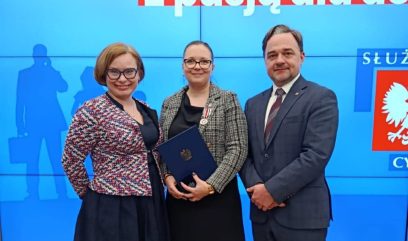 Trzy dorosłe osoby w oficjalnych strojach stoją uśmiechnięte na niebieskim tle z polskim tekstem. Kobieta w centrum trzyma teczkę i nosi medal. Druga kobieta nosi okulary i wzorzystą marynarkę; mężczyzna obok nich ma na sobie garnitur i krawat.