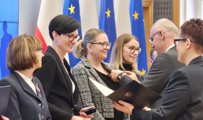 Grupa ludzi w formalnych strojach stoi w pomieszczeniu. Mężczyzna wręcza certyfikat i przypina odznakę uśmiechniętej kobiecie. W pobliżu stoją trzy inne kobiety. Za nimi znajdują się flagi Unii Europejskiej i Polski. Atmosfera wydaje się być ceremonią wręczenia nagrody lub uznania.