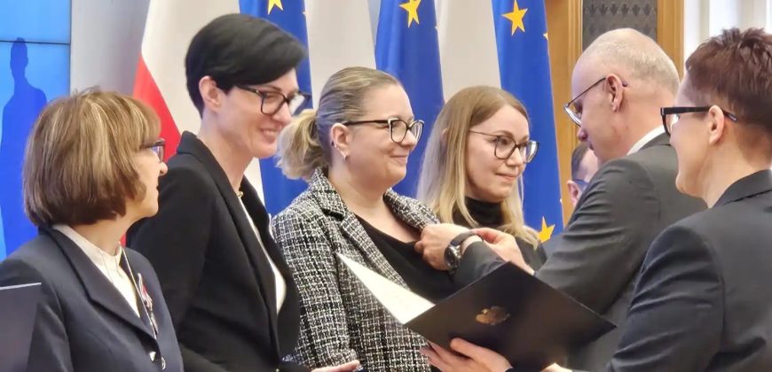 Grupa ludzi w formalnych strojach stoi w pomieszczeniu. Mężczyzna wręcza certyfikat i przypina odznakę uśmiechniętej kobiecie. W pobliżu stoją trzy inne kobiety. Za nimi znajdują się flagi Unii Europejskiej i Polski. Atmosfera wydaje się być ceremonią wręczenia nagrody lub uznania.