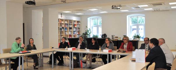 Grupa ludzi w strojach biznesowych siedzi przy stołach ustawionych w kształt litery U. W tle okna i regał z książkami