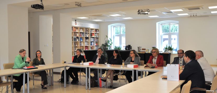 Grupa ludzi w strojach biznesowych siedzi przy stołach ustawionych w kształt litery U. W tle okna i regał z książkami