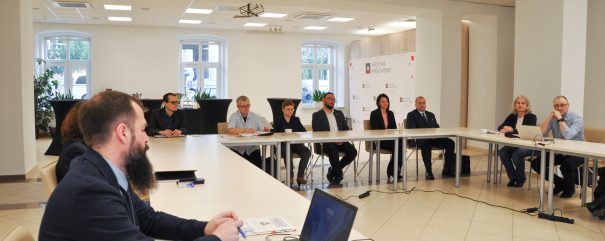 Grupa ludzi siedzi wokół stołów w kształcie litery U w jasnej sali konferencyjnej, słuchając mężczyzny z laptopem i dokumentami na pierwszym planie. Duże okna i białe ściany tworzą otwartą atmosferę, a w tle stoi wyświetlacz prezentacji.