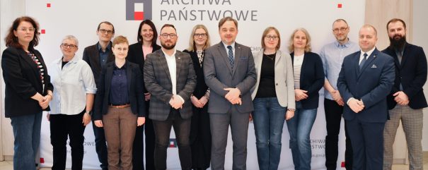 Grupa piętnastu osób, zarówno mężczyzn, jak i kobiet, stoi w pomieszczeniu przed białym tłem z logo i nazwą 