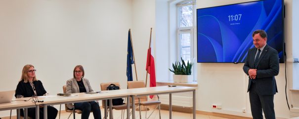 Mężczyzna w garniturze stoi w pobliżu ekranu wyświetlającego godzinę 11:07 w jasnej sali konferencyjnej. Dwie kobiety siedzą przy stole konferencyjnym z laptopami. Przy oknie widoczne są flagi Polski i UE. Pokój ma białe ściany, zegar i rośliny doniczkowe.