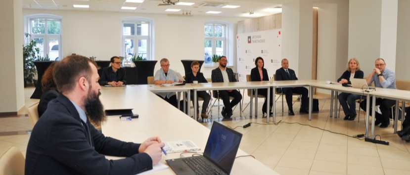 Dwanaście osób siedzi wokół stołów w kształcie litery U w jasnej sali konferencyjnej. Niektórzy robią notatki lub uważnie słuchają. Na pierwszym planie widoczny jest laptop i dokumenty. W tle pojawiają się duże okna i biała tablica prezentacyjna.