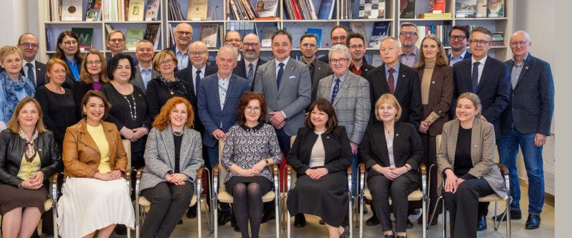 Grupa około 30 dorosłych osób, zarówno mężczyzn, jak i kobiet, w tym Kierownictwo, pozuje razem przed regałami wypełnionymi książkami i dokumentami. Sceneria wydaje się być oficjalną lub akademicką biblioteką z państwowej sieci archiwalnej.