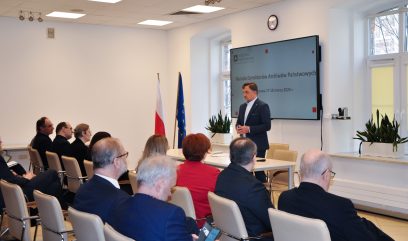 Mężczyzna w garniturze przemawia z podium do siedzącej publiczności w sali konferencyjnej. Za nim znajdują się flagi Polski i UE, rośliny doniczkowe, duże okna i ekran wyświetlający prezentację w języku polskim. Większość słuchaczy jest ubrana formalnie.