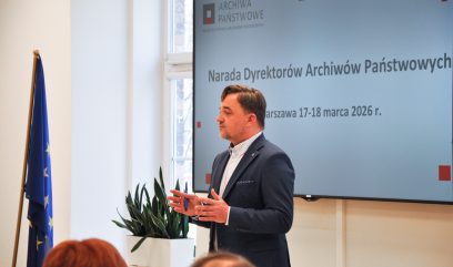 Mężczyzna w garniturze stoi i przemawia przed ekranem prezentacyjnym wyświetlającym polski tekst o spotkaniu dyrektorów w marcu 2026 roku. W pobliżu widoczna jest flaga UE i zielona roślina. Ludzie siedzą naprzeciw niego, słuchając uważnie.