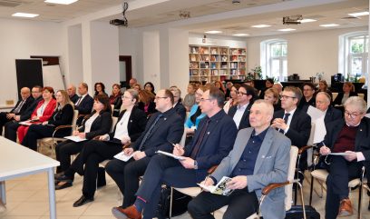 Grupa mężczyzn i kobiet, w większości ubranych w stroje biznesowe, siedzi uważnie w rzędach krzeseł w jasnym, nowoczesnym pokoju z regałami i dużymi oknami, słuchając prezentacji lub wykładu. Niektórzy robią notatki, podczas gdy inni trzymają broszury.