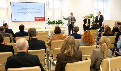 Mężczyzna w szarym garniturze gestykuluje podczas prezentacji dla publiczności siedzącej w jasnej sali konferencyjnej. Dwóch kolegów stoi obok niego. Za nimi duży ekran wyświetla polską prezentację. Uczestnicy są zwróceni do przodu, słuchając uważnie, a przy oknach stoją rośliny doniczkowe.