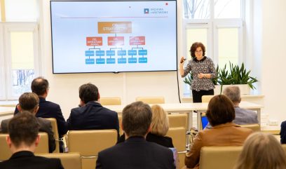 Kobieta prowadzi prezentację dla siedzącej publiczności w sali konferencyjnej. Stoi przy dużym ekranie wyświetlającym diagram z polskim tekstem, w tym CEL STRATEGICZNY. Uczestnicy zwróceni są twarzami do przodu, a przez okna wpada światło dzienne.