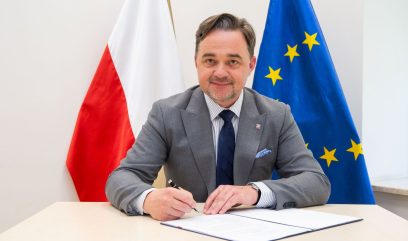 Mężczyzna w szarym garniturze siedzi przy biurku i podpisuje dokument. Za nim znajduje się flaga Polski (biało-czerwona) i flaga Unii Europejskiej (niebieska z żółtymi gwiazdami). Mężczyzna patrzy w kamerę i lekko się uśmiecha.