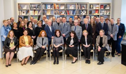 Grupa około 35 dorosłych osób, mężczyzn i kobiet, pozuje razem w pomieszczeniu przed regałami wypełnionymi książkami i dokumentami. Większość stoi, a siedem kobiet siedzi z przodu. Otoczenie wydaje się formalne i profesjonalne.