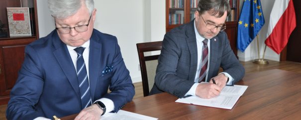 Dwóch mężczyzn w garniturach siedzi przy drewnianym stole i podpisuje dokumenty. W tle widoczne są półki z książkami i zegar.