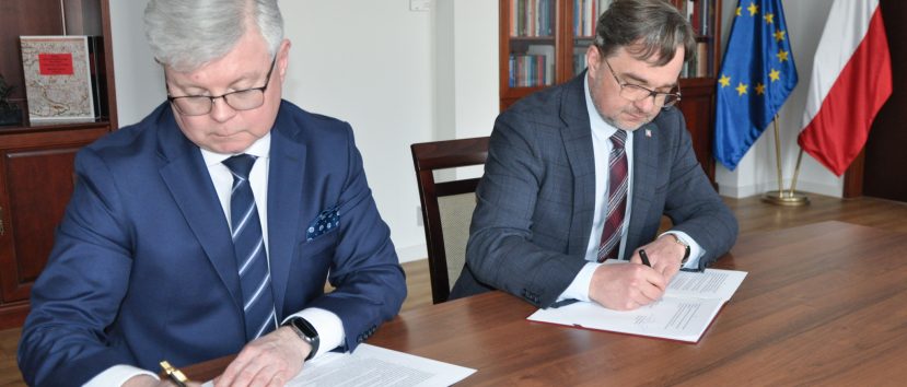 Dwóch mężczyzn w garniturach siedzi przy drewnianym stole i podpisuje dokumenty. W tle widoczne są półki z książkami i zegar.