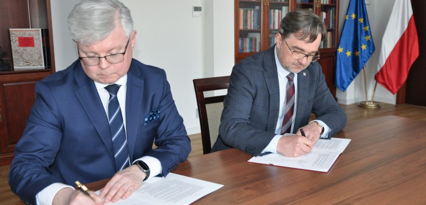 Dwóch mężczyzn w garniturach siedzi przy drewnianym stole i podpisuje dokumenty. W tle widoczne są półki z książkami i zegar.