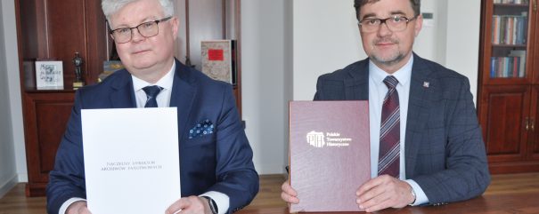 Dwóch mężczyzn w garniturach siedzi przy drewnianym stole, a każdy z nich trzyma dużą teczkę. Mężczyzna po lewej stronie trzyma białą teczkę, a mężczyzna po prawej brązową teczkę z białym logo. W tle widoczne są półki z książkami i zegar.