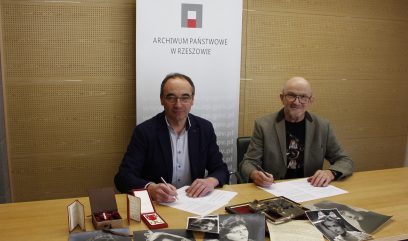 Dwóch mężczyzn w garniturach siedzi przy stole podpisując dokumenty, na którym rozłożone są zabytkowe czarno-białe zdjęcia i medale. Za nimi znajduje się baner z napisem Archiwum Państwowe w Rzeszowie, sugerujący kontekst archiwalny lub historyczny.