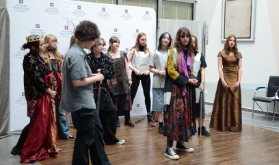 Grupa młodych ludzi stoi w półkolu na drewnianej podłodze w pomieszczeniu, ubrana w eklektyczne, artystyczne stroje. Na ścianie za nimi wisi białe tło z logo i oprawiony szkic. Niektórzy patrzą w kamerę, inni patrzą gdzie indziej.