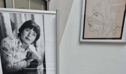 Czarno-białe zdjęcie kobiety z krótkimi ciemnymi włosami i wzorzystą koszulą, opierającej się na jednym ramieniu, obok oprawionego w ramkę rysunku kobiecego profilu na białej ścianie. Oba obrazy są wyświetlane w wewnętrznej przestrzeni wystawienniczej.