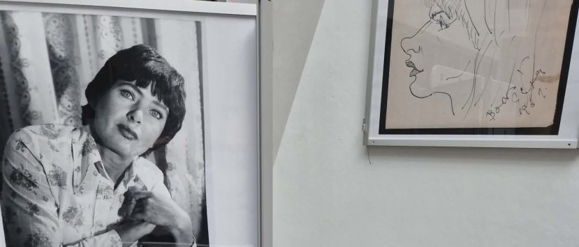 Czarno-białe zdjęcie kobiety z krótkimi ciemnymi włosami i wzorzystą koszulą, opierającej się na jednym ramieniu, obok oprawionego w ramkę rysunku kobiecego profilu na białej ścianie. Oba obrazy są wyświetlane w wewnętrznej przestrzeni wystawienniczej.