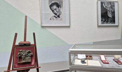 Oprawiona abstrakcyjna płaskorzeźba stoi na drewnianej sztaludze obok szklanej gabloty z papierami i nagrodami. Dwa duże czarno-białe portrety kobiet wiszą na bladej ścianie z geometrycznymi wzorami w tle.