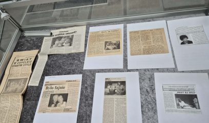 Na szarej powierzchni pod szklaną gablotą znajduje się kilka starych wycinków z gazet i artykułów w języku polskim. Niektóre zawierają czarno-białe zdjęcia ludzi i scen. Gazety różnią się rozmiarem, a większość tekstów wydrukowano w kolumnach.