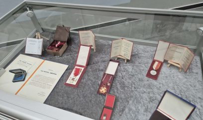 Szklana gablota prezentuje różne medale, certyfikaty i nagrody na szarej powierzchni. Przedmioty obejmują zapakowane w pudełka medale, certyfikaty umieszczone na małych sztalugach i oficjalnie wyglądające dokumenty, wszystkie ułożone schludnie w muzeum lub na wystawie.