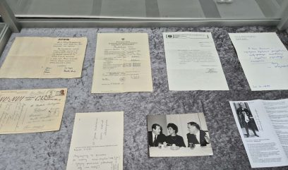 Pokazano gablotę z ośmioma historycznymi dokumentami i fotografiami. Przedmioty obejmują listy pisane na maszynie i odręcznie, pocztówkę, czarno-białe zdjęcie trzech osób rozmawiających przy stole i małe zdjęcie stojącej osoby, wszystkie ułożone na szarej filcowej powierzchni.